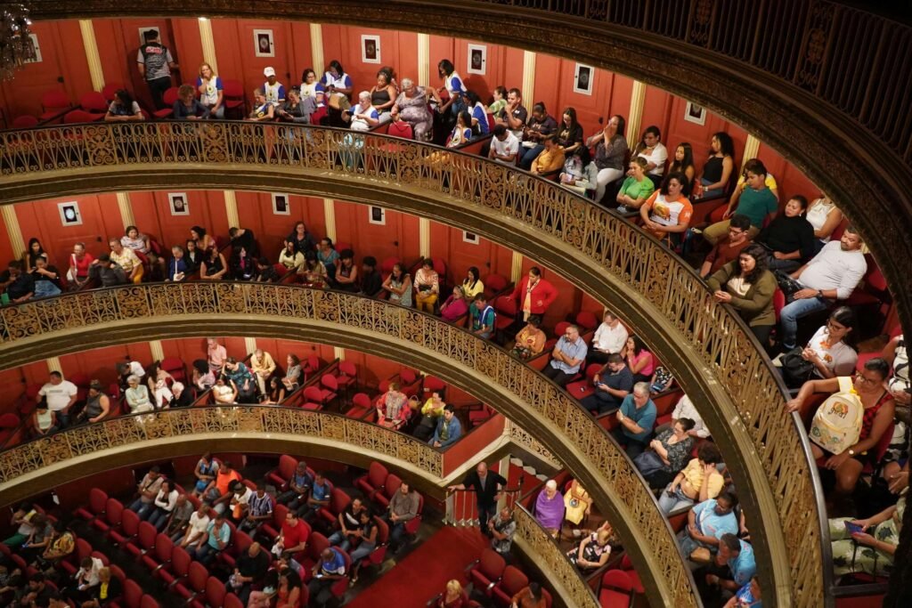 Packed audience seated across multiple ornate balcony tiers inside Teatro Nacional, Costa Rica, with gold ironwork railings and red seating