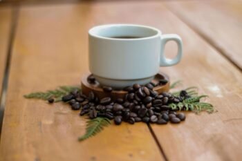 White ceramic cup of freshly brewed Costa Rican coffee surrounded by roasted coffee beans and green fern leaves on a wooden table at a coffee farm