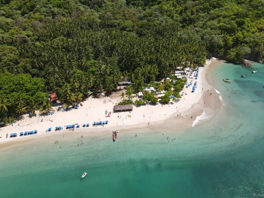 Aerial view of a palm-backed beach lined with blue umbrellas, kayaks, and boats anchored in a turquoise cove