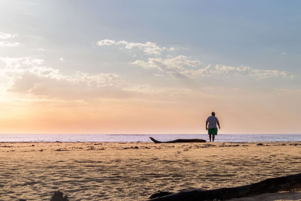 Person standing at the shoreline at sunset on a wide, empty beach with driftwood on the sand, similar to a quiet Costa Rica beach