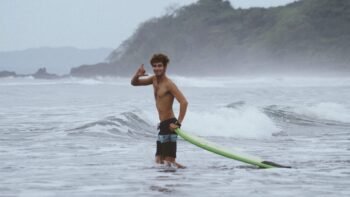 Smiling surfer throwing a shaka while holding a green longboard in the waves, similar to a Costa Rica surf break
