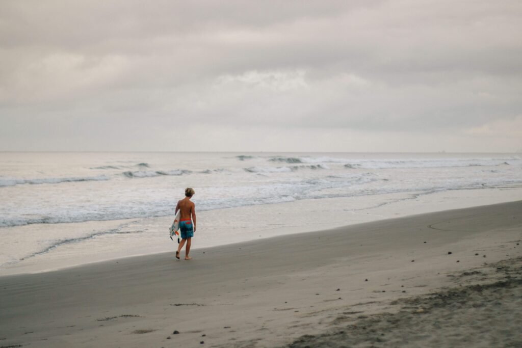 Surfer walking along an empty dark-sand beach carrying a board under an overcast sky, similar to a quiet Costa Rica surf beach