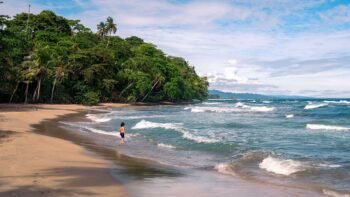 Child wading in shallow surf along a quiet, palm-fringed beach below a jungle headland, similar to Costa Rica's Pacific coast