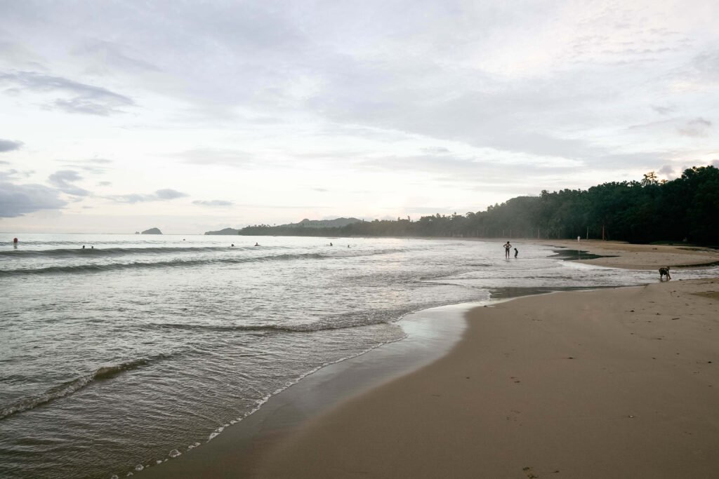 Calm shallow water and wide sand at dawn with a few swimmers and a forested headland, similar to Playa Cabuyal, Costa Rica