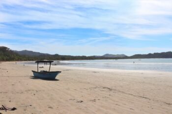 Wide pale sand beach at low tide with a small beached boat and dry-forest hills behind, similar to Playa Cabuyal, Costa Rica
