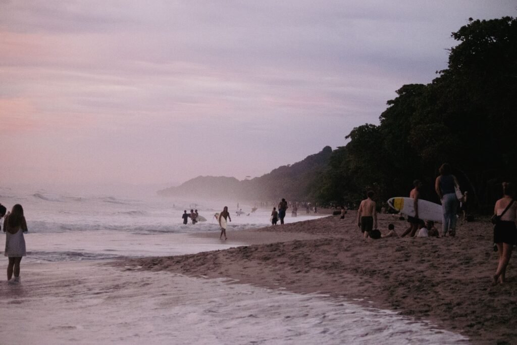Surfers and beachgoers along a Pacific beach at dusk below a forested headland, similar to Playa Hermosa, Costa Rica